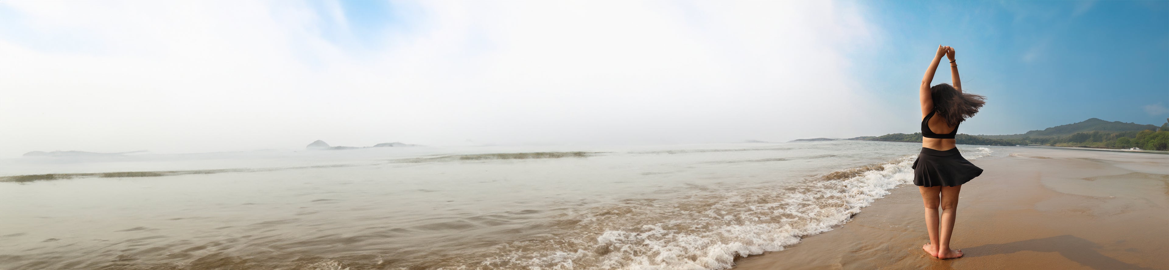 Woman in a black dress standing on a beach with ocean waves and clear sky.